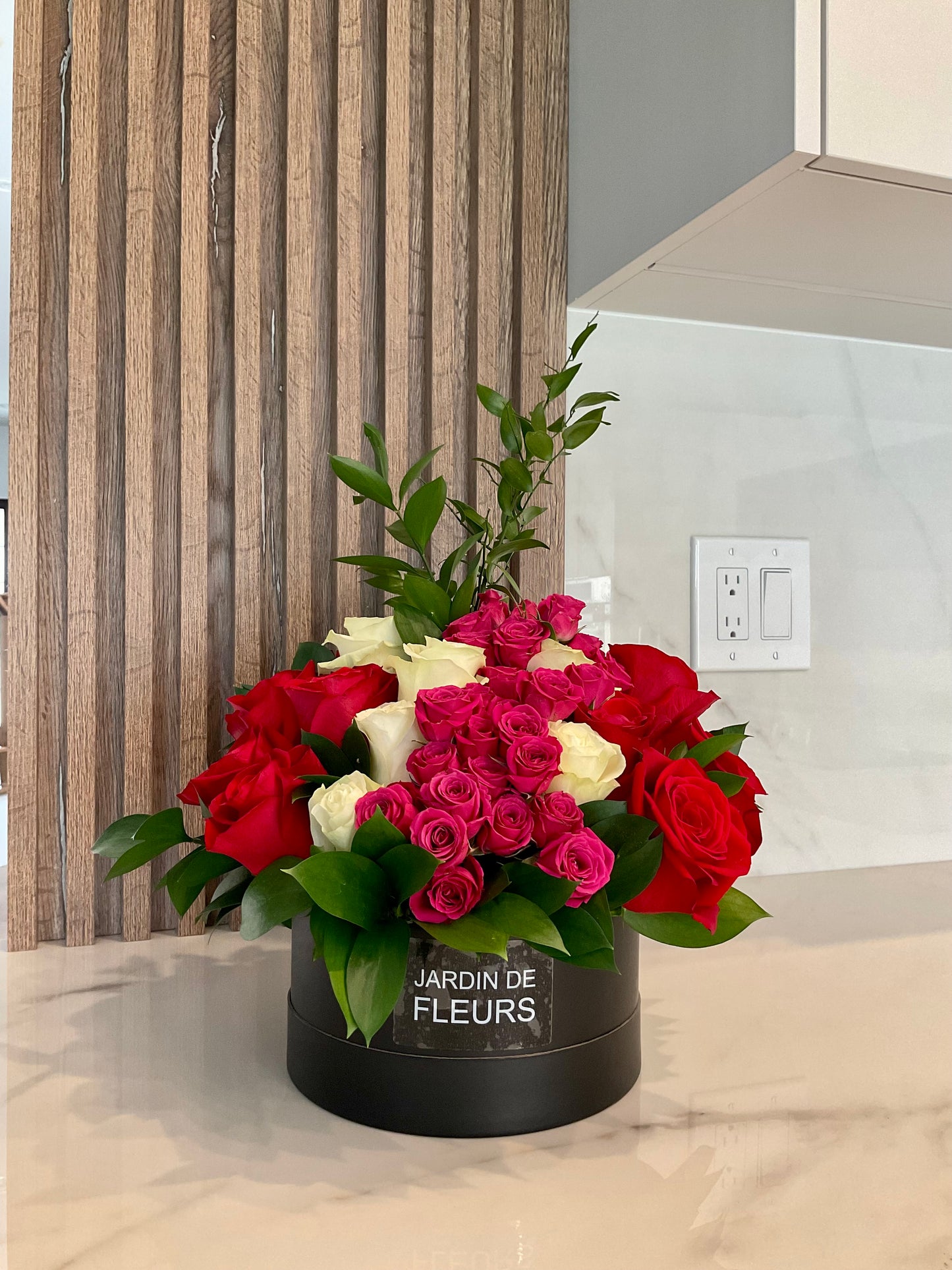 Bouquet of red, pink, and white roses in a black container labeled 'Jardin de Fleurs' on a kitchen floor.