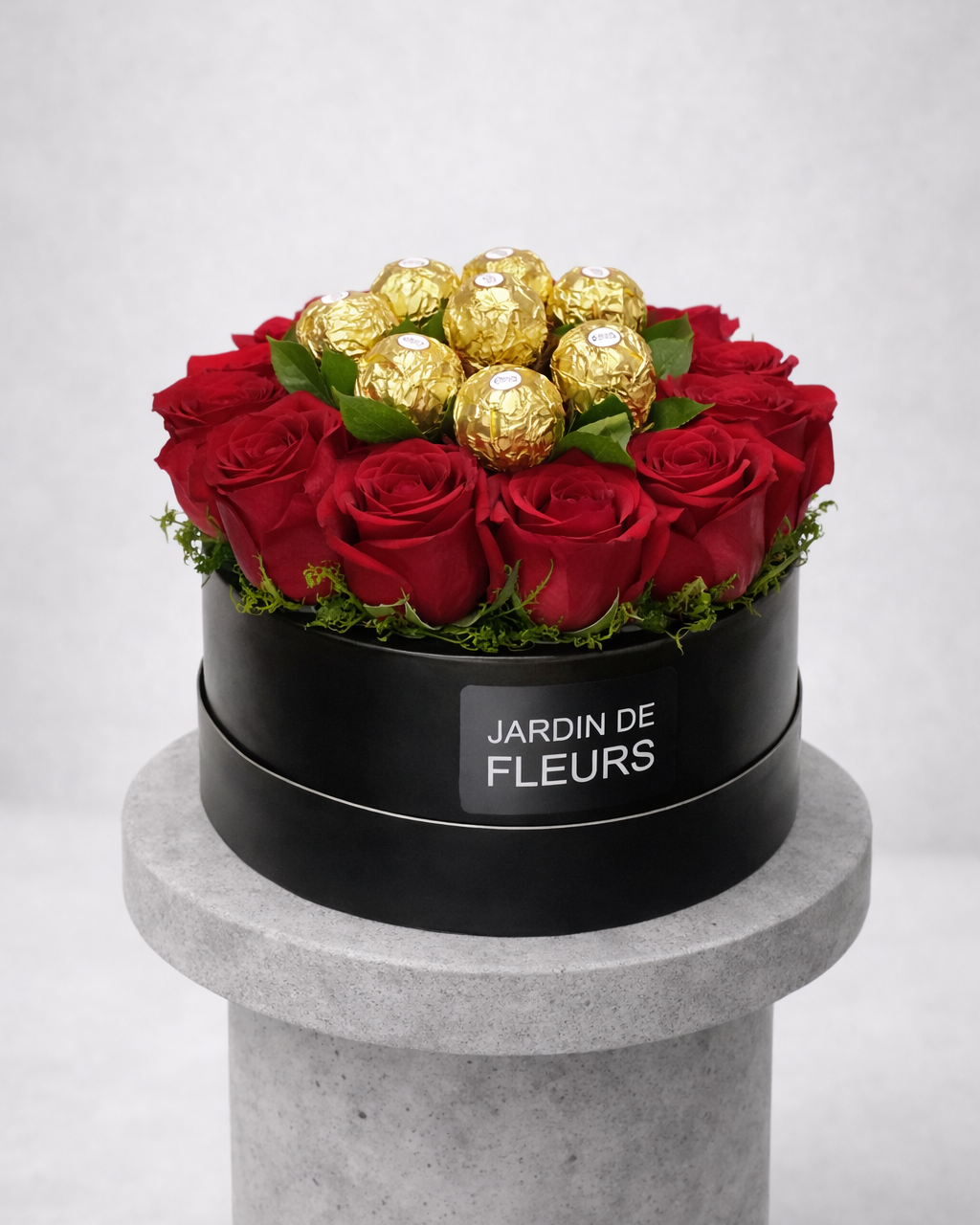 Black box of Jardin de Fleurs with red roses and gold Ferrero Rocher chocolates on a gray pedestal.