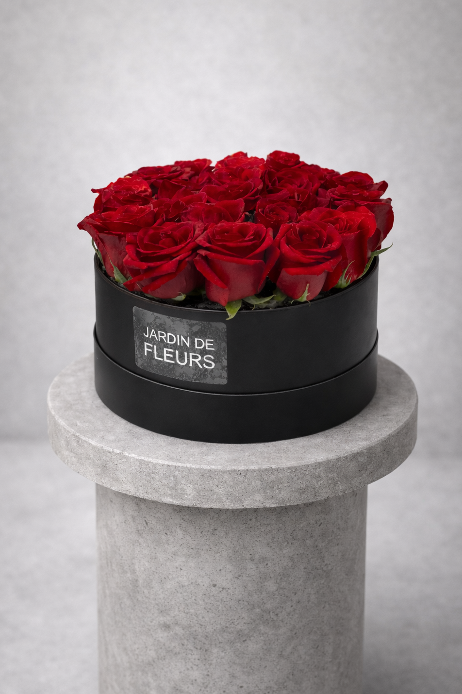 Black round box with red roses labeled 'Jardin de Fleurs' on a gray pedestal against a light gray background