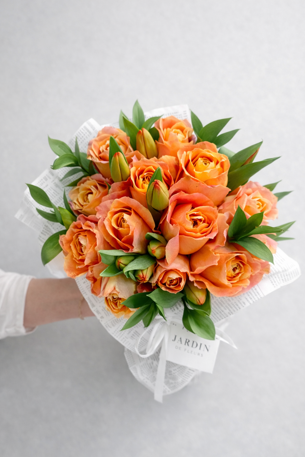 Bouquet of orange roses with green leaves held by a person on a light gray background, with 'JARDIN' branding visible.